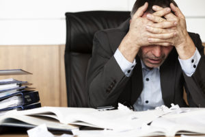 A businessman sitting in front of a cluttered desk with his hands crossed over his face in frustration.