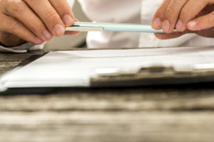 Two hands holding a pen parallel over a clip board.
