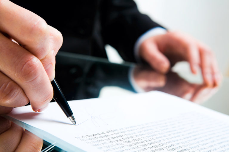 Two hands on a table, one holding a pen and signing a paper document.