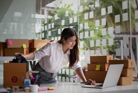 A business woman standing over a desk with a laptop and a wall of sticky notes behind her.