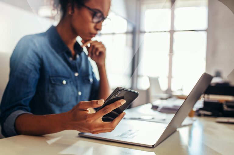 a woman sitting at a desk, looking at a laptop while holding a cell phone